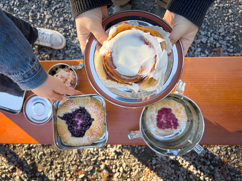 Young woman holding a cinnamon bun in a stainless steel container while another picks up a blueberry danish from a picnic bench.  Vancouver, British Columbia, Canada.