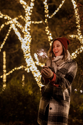Portrait of cheerful young woman standing at the park, against glistening Christmas lights, waving a sparkler, smiling and making a wish.