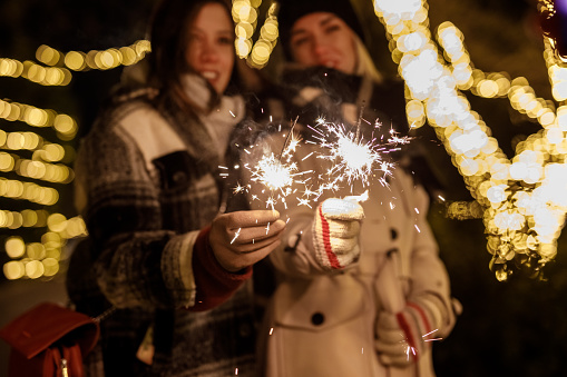 Selective focus shot of two joyful girlfriends standing at the park decorated with glistening Christmas lights, having bonding moments while waving sparklers.