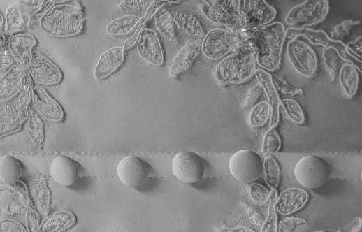 Black and white photo of a section of an old white wedding dress, showing 6 covered buttons on a seam and flowery embroidery with a pearl in the centre of the flower