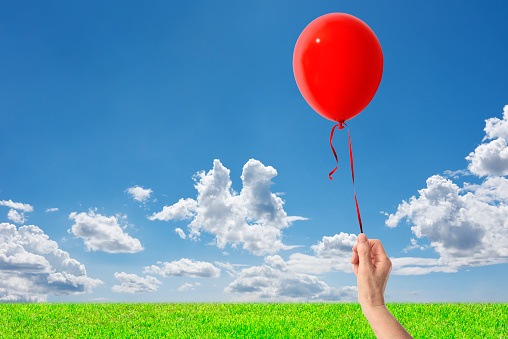 Hand holding a red balloon in the blue sky over the green hill with copy space.