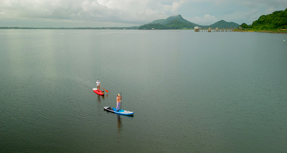 Aerial view of Asian athletic woman on paddle board with friend at lake. Outdoor water sport and travel on summer holiday Thailand.