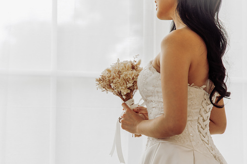 A side view of a bride holding a delicate bouquet, the intricate details of her wedding gown's bodice highlighted by the soft light streaming through sheer curtains