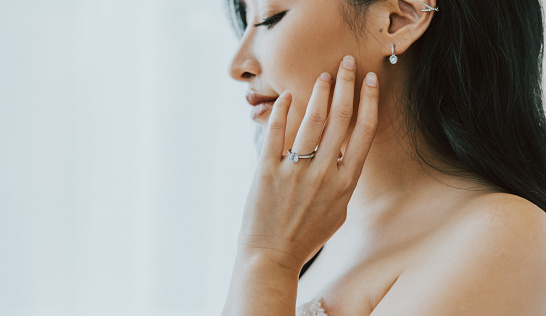 A bride in contemplative repose, her hand gracing her cheek, showcasing the sparkle of her engagement ring and the elegance of her earrings in a soft, natural light