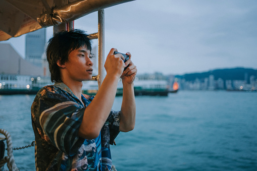 Young Asian man looking out through the window taking photos while traveling by ferry at Victoria Harbour
