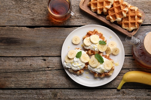 Delicious Belgian waffles with banana and whipped cream served on wooden table, flat lay. Space for text