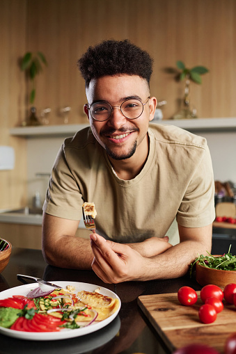 Young middle eastern influencer looking at camera holding fork with piece of omelet leaning on kitchen table with dish and vegetables on it