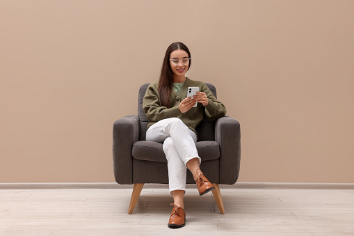 Beautiful woman with smartphone sitting in armchair near beige wall indoors