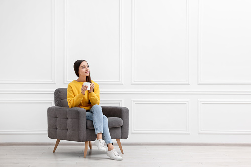 Beautiful woman with cup of drink sitting in armchair near white wall indoors, space for text