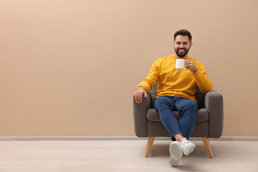 Handsome man with cup of drink sitting in armchair near beige wall indoors, space for text