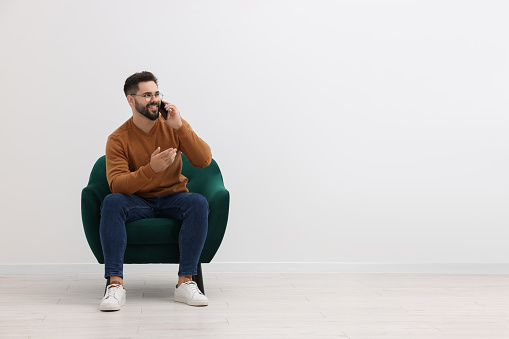 Handsome man talking on smartphone while sitting in armchair near white wall indoors, space for text