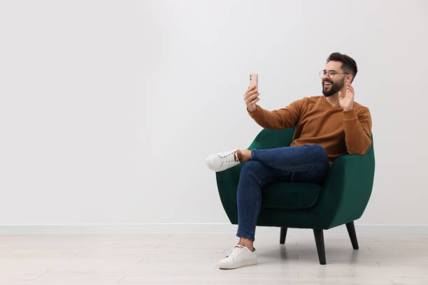 istock Handsome man having video chat via smartphone while sitting in armchair near white wall indoors, space for text 1831900140