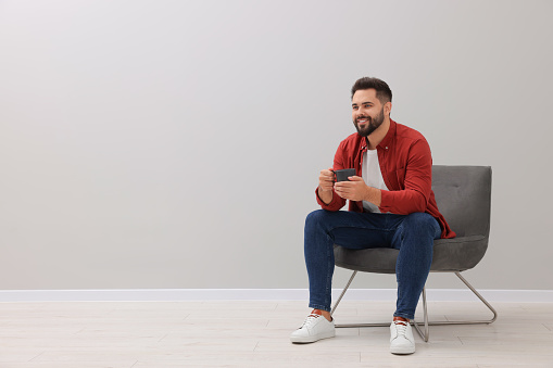 Handsome man with cup of drink sitting in armchair near light grey wall indoors, space for text