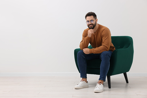 Handsome man sitting in armchair near white wall indoors, space for text