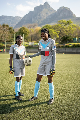 Portrait of two girls in sports clothing with a soccer ball standing together on a sports field outdoors looking at camera and smiling
