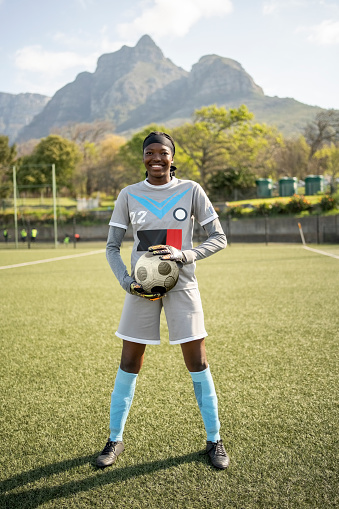 Ful length portrait of confident young soccer girl holding ball standing on field looking at camera and smiling outdoors
