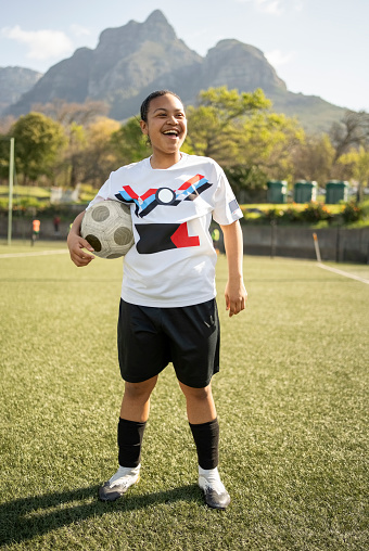 Full length portrait of cheerful young soccer player holding ball standing on field looking at away and smiling outdoors