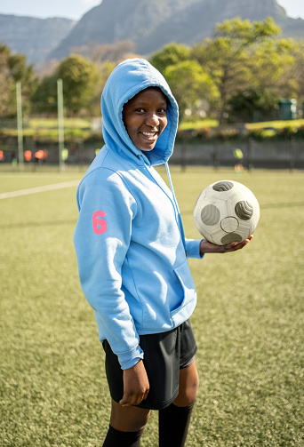 Portrait of happy young female soccer player in sports hoodie standing with a soccer ball on field outdoors
