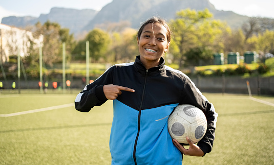 Portrait of a smiling teenage girl in sportswear pointing her finger at soccer ball in her hand while standing on outdoor sports field