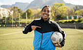 Portrait of happy teenage girl pointing at soccer ball standing on outdoor sports field
