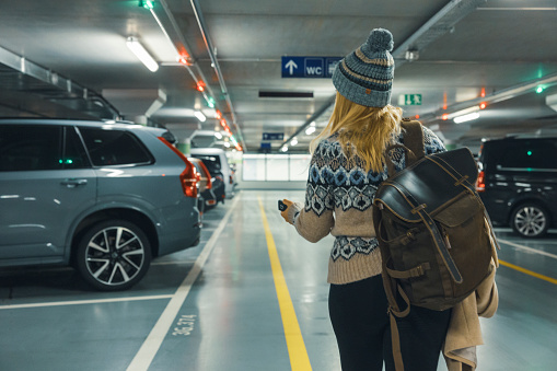 Rear view of a woman traveler in warm clothing carrying backpack in the parking lot unlocking her car