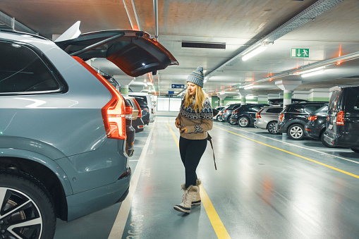 Woman traveler in warm clothing about to load her bag in trunk of her car at underground parking lot