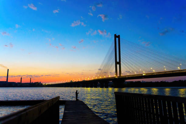 Silhouette of a fisherman against the backdrop of the Southern Bridge over the Dnipro River at sunset. Travel, fishing and sunset-theme related background.