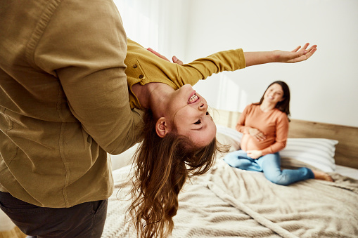 Cheerful girl having fun with her father in bedroom. Mother is pregnant and relaxing in the background. Copy space.