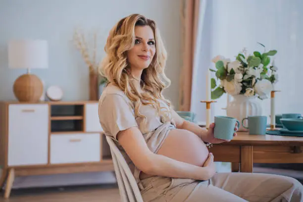 Happy pregnant young blonde caucasian woman sitting at table touching belly feels baby moving inside, female dreaming about newborn baby at home. Healthy pregnancy.