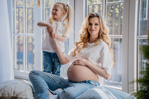 Blonde pregnant young woman in jeans playing with little daughter at home sitting on floor on sunny summer day. Healthy family.