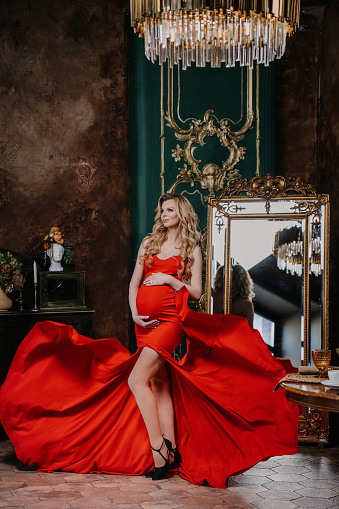 Vertical shoot of blonde caucasian pregnant woman in red dress looks away standing at antique interior. Pregnancy, healthy women. Classical photoshoot.