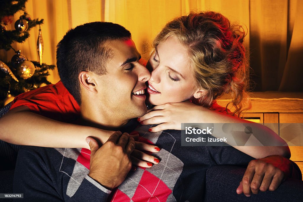 Romantic Christmas couple Happy young adult couple celebrating Christmas. Woman embracing her partner and kissing him.  Christmas tree in the background. Blond Hair Stock Photo