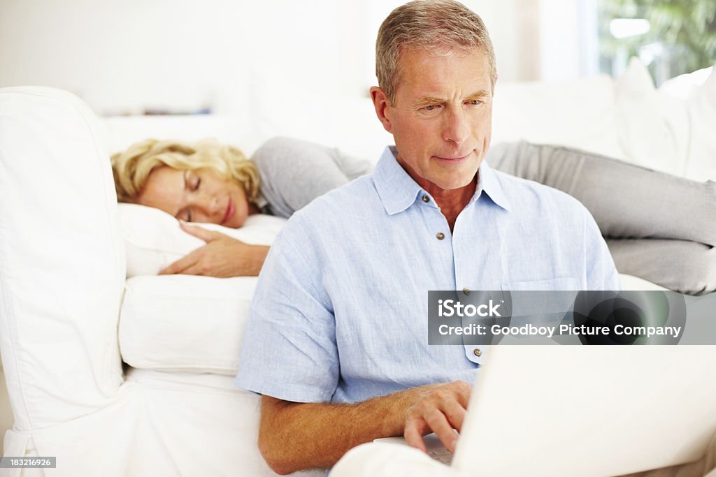 Homem sênior trabalhando no laptop com mulher dormindo ao fundo - Foto de stock de 40-49 anos royalty-free