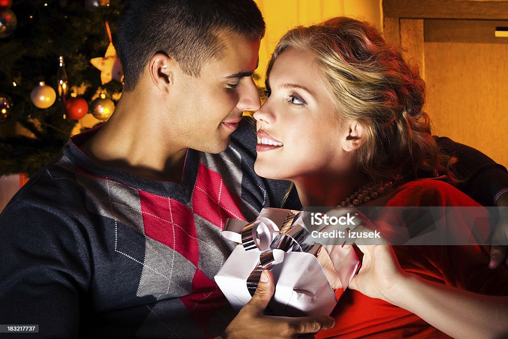 Christmas Gifts "Happy young adult couple exchanging Christmas gifts. They holding white gift box with silver ribbon, embracing and looking eatch other's eyes. Christmas tree in the background." Blond Hair Stock Photo