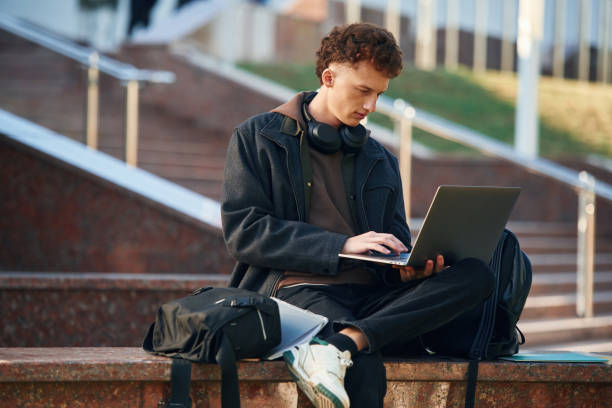 istock Guy is sitting and using laptop. Young student is outside the university, in black clothes 1832221675