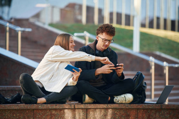 istock Holding smartphone. Two young students are sitting outdoors against university 1832223800