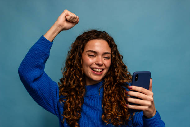 Happy young woman in blue sweater using smartphone and making winner gesture on blue background