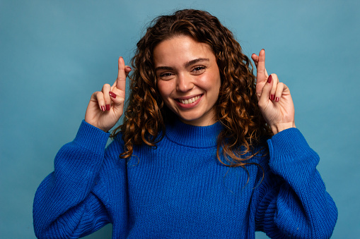 Smiling girl in a blue sweater on a blue background with fingers crossed