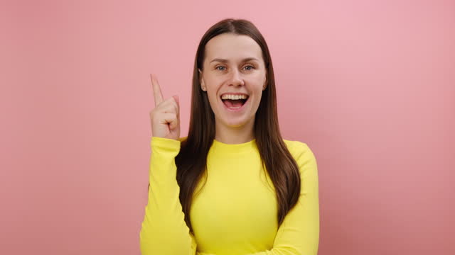 Overjoyed happy cute young woman pointing finger up looking at camera with toothy smile, having idea, startup, wearing yellow sweater, isolated on pink color background wall in studio. Plan concept