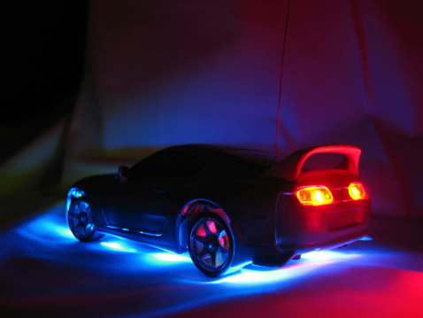 A macro shot of the rear of a remote control model street racer car in the dark with blue neon illumination under the body and red taillights.  Low noise thanks to Neat Image.  Focus is sharp at the rear wheel and softens at the front.More in the series: