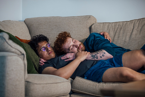 Close-up shot of a gay couple relaxing at home. They are embracing each other asleep on the sofa. They are both lying comfortably wearing cozy clothing on the sofa at home. Located in Durham, England.