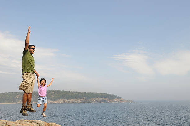 A father and daughter wearing hiking attire jump in the air along a rocky coastline.