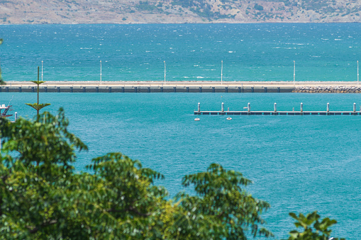 Green leaves of trees against the background of turquoise or blue ocean, sea, in the middle ground the shoreline intended to dampen the waves entering the pier, shore in background.