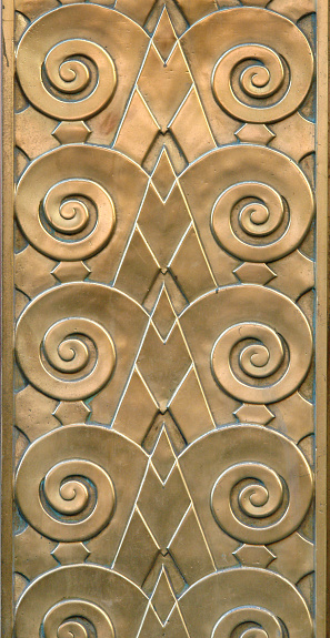 Detail of an Art Deco style bronze relief. The photo was taken in New york City and features a repeating pattern juxaposing strong rising diagonal elements with spiral curlicues suggesting rams horns. The bronze has a rich array of golden hues and is embued with a patination conducive to a structure built in 1929. (See more in the \