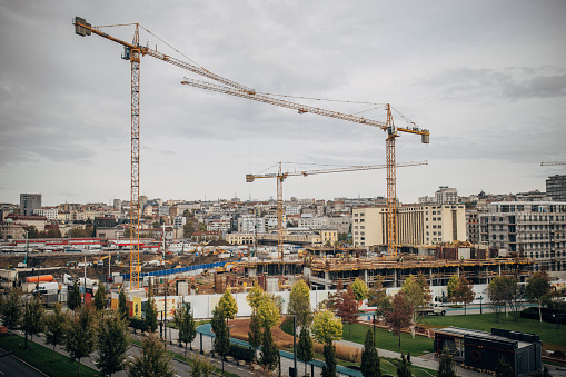A view of a construction site in the city with modern buildings and cranes in the background surrounded by a green fence and trees during an overcast day.