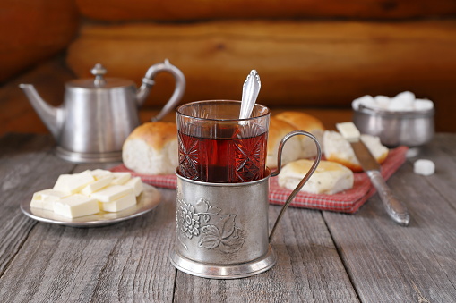 On a wooden background are a glass of hot tea in a metal cup holder in front of delicious buns on a red textile, a metal plate with butter and other utensils.