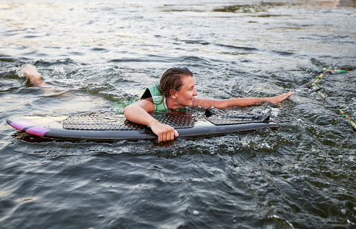 Young beautiful sporty girl in a green life jacket swims in the water with a wakeboard in her hand. Happy sportswoman is preparing for a steep wakeboard ride. Active lifestyle, healthy hobby.