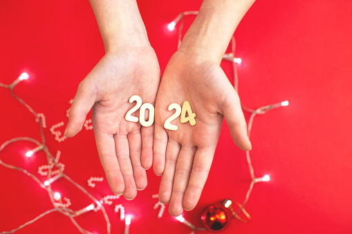 Top view of child hands holding wooden number 2024 on palms on red garland lights background with copy space. Christmas greeting card, New Year, winter holidays sale and festive mood concept.