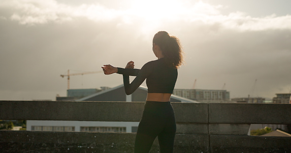 Woman, stretching and fitness in city for workout, exercise or outdoor training in sunset. Rear view of female person, runner or athlete in body warm up or arm stretch getting ready in an urban town