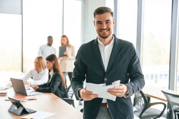 istock White man in suit is with papers in hands, standing. Group of office workers are together indoors 1832440264